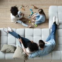 Above view parents sit on comfortable sofa while multi-ethnic daughters play on carpet using bricks set and dinosaur toys, new home and furniture, modern house, rented apartment family weekend concept