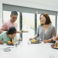 Family are having dinner together at home. The little girl is struggling with a too-big mouthful of noodles and her family are watching her, laughing.